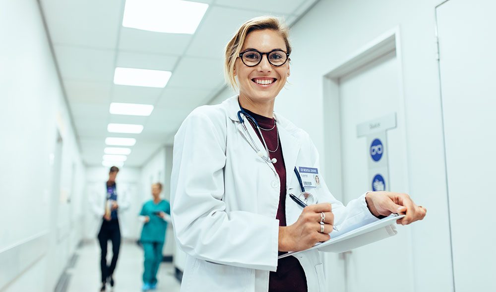 A physician walking a hospital corridor, smiling, with a clipboard in hand.